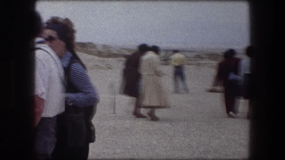 1976 Messada, Israel home-movie frame showing beach visitors in coats and casual clothing near a stone-lined shore.
