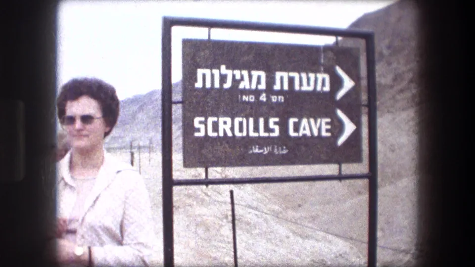 A woman in a light tan coat and sunglasses stands beside the Scrolls Cave sign at Masada in 1976.