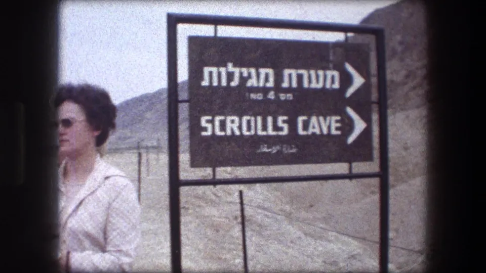 1976 Masada home-movie frame with a woman partly in view beside the Scrolls Cave sign and desert hills.