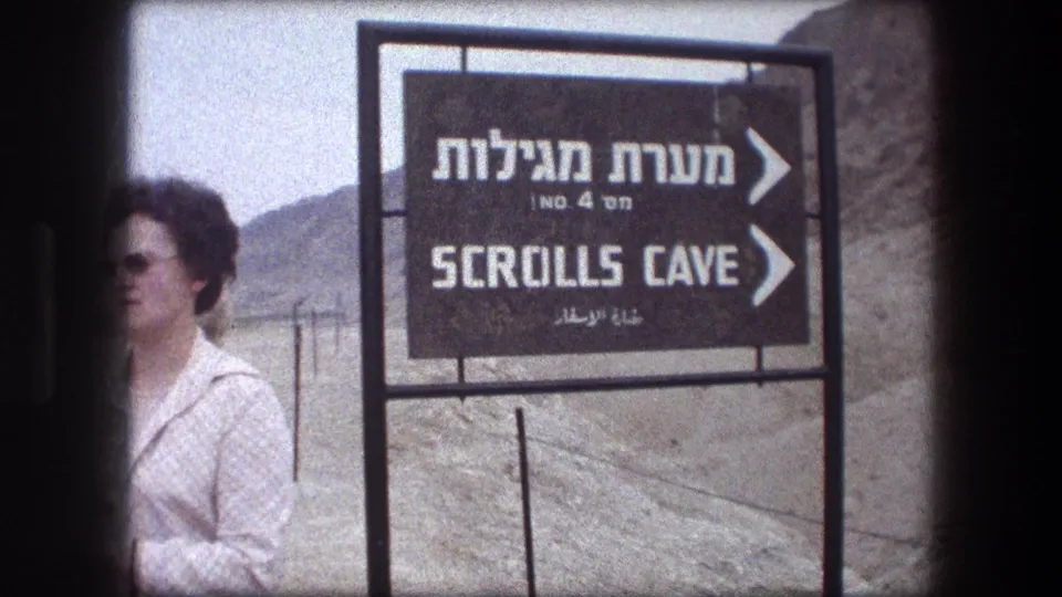 1976 8mm frame of a tourist near a Hebrew, English, and Arabic Scrolls Cave sign at the Qumran site in Israel.