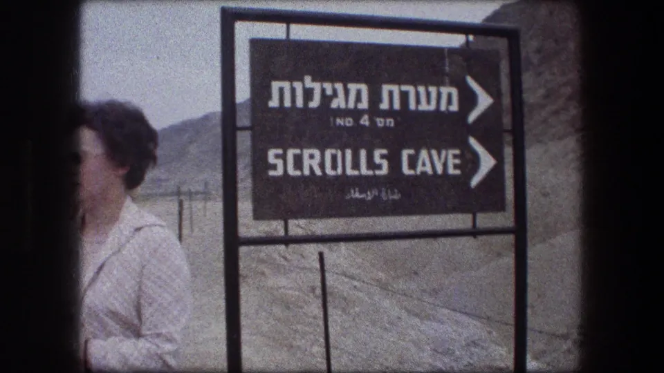 Close 8mm view of the Qumran Scrolls Cave direction sign with Hebrew, English, and Arabic lettering and a partially visible tourist.