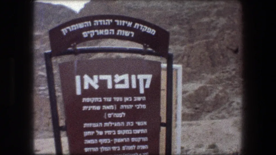 1976 8mm frame showing a Hebrew Qumran site plaque with arid hills and metal supports in the background.