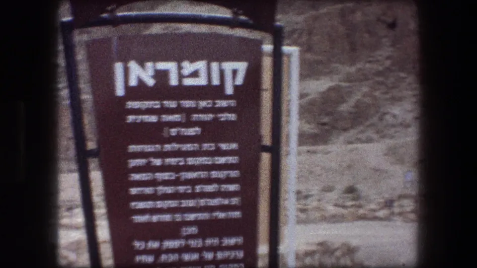 Close 8mm view of a Qumran informational plaque with Hebrew text, metal framing, and dry terrain behind it.