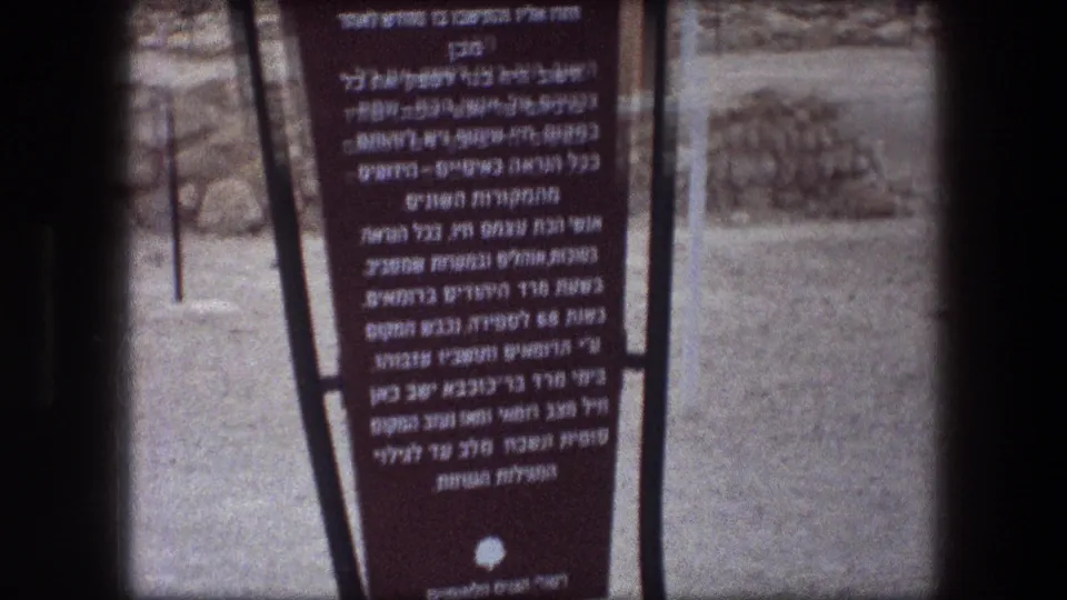 1976 8mm frame showing the lower portion of a Hebrew informational sign at Messada against rocky arid terrain.