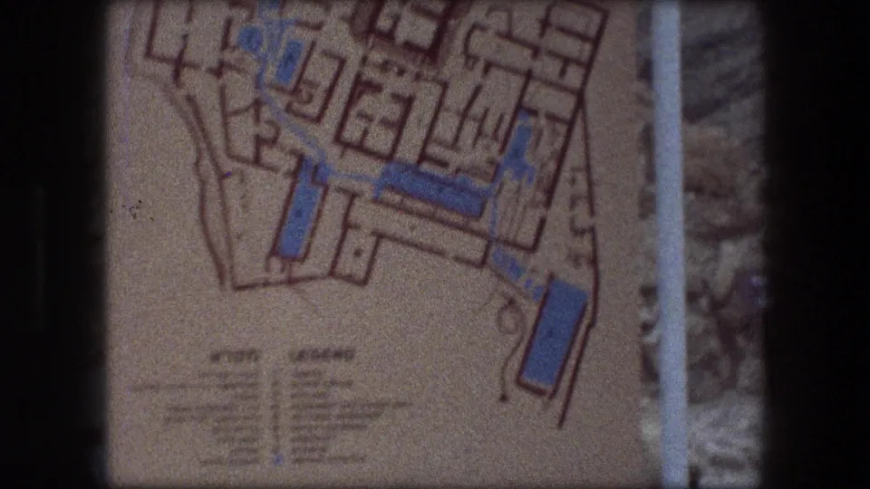 Final 1976 Messada frame holding on the printed site-plan sign, with blue features, orange outlines, and a small legend below.