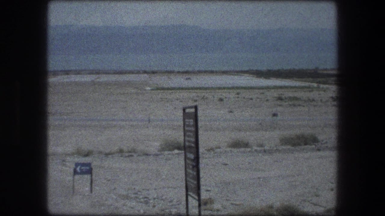 Desert landscape with a tall signpost, smaller blue sign, sparse scrub, and distant hills.