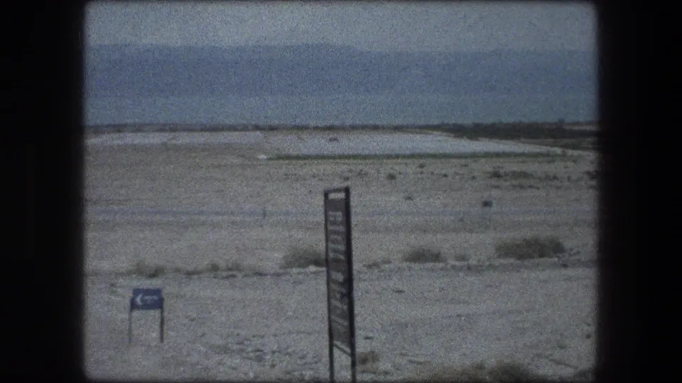 1976 8mm view of Messada, Israel desert terrain with foreground signage, pale ground, and a hazy horizon.