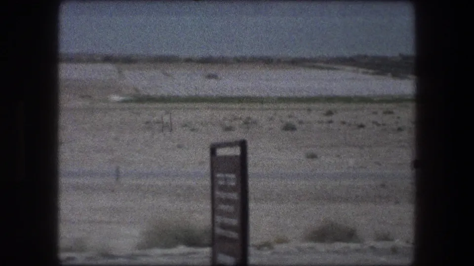 1976 8mm frame of Messada, Israel showing a signpost at the edge of a barren desert plain under a hazy sky.
