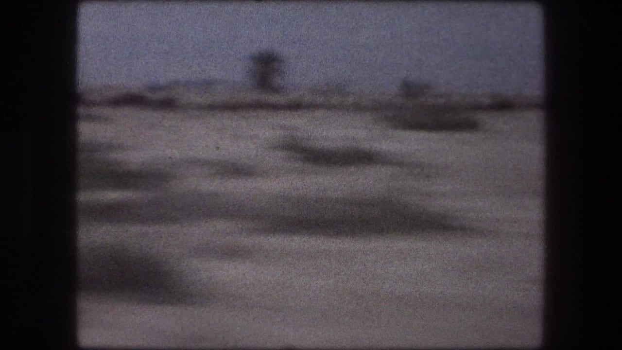 Train-window frame from 1976 Israel footage, with sparse terrain and indistinct distant forms sliding past.