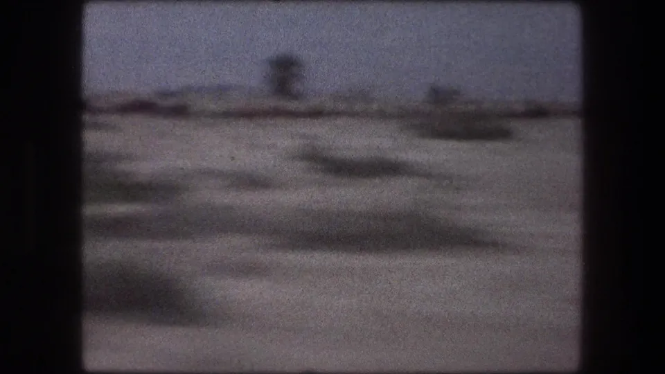 Train-window frame from 1976 Israel footage, with sparse terrain and indistinct distant forms sliding past.