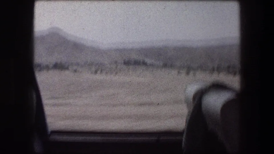 1976 Messada-area train view with a distant hill, sparse vegetation, and part of the carriage interior at the edge.