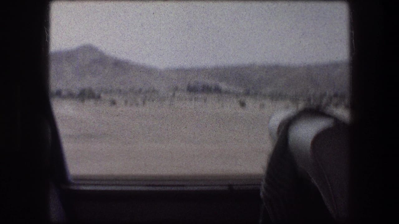 Desert landscape and distant hills seen through a vehicle window with a seat back at right.