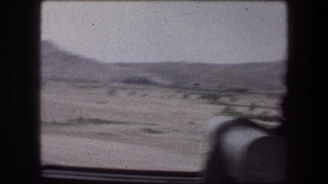Vehicle-window view across a dry landscape in the Messada travel sequence, with low hills, pale sky, and interior seat framing the scene.