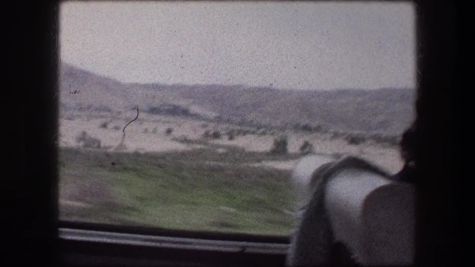 1976 8mm passenger view showing green roadside patches, pale desert flats, and distant hills through a vehicle window near Messada.