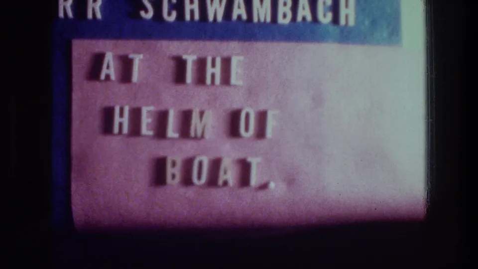 1970s 8mm title card with the phrase “AT THE HELM OF BOAT.” and a partially visible name line at the top.