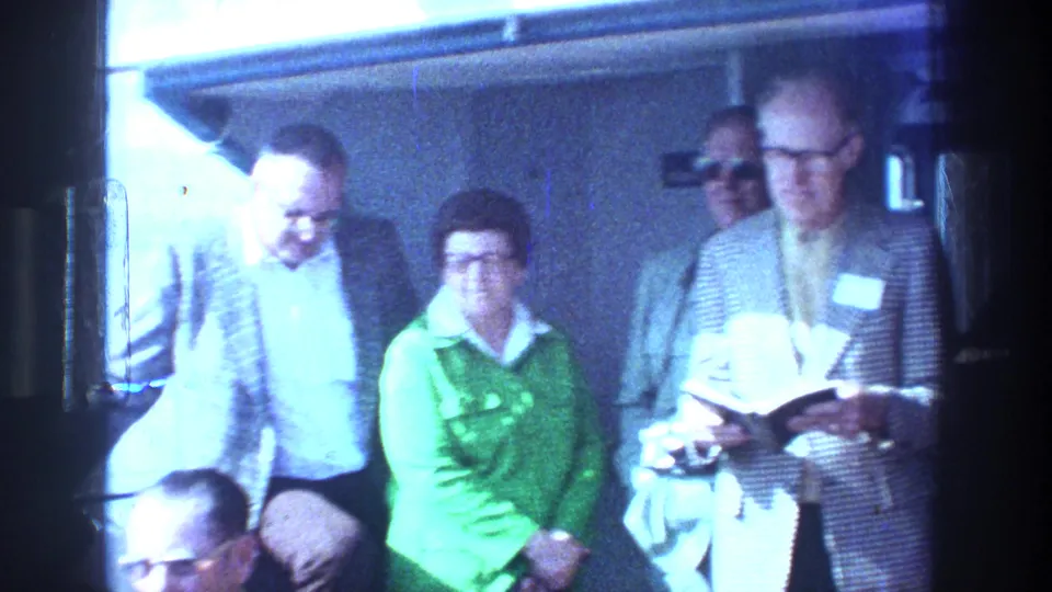 Aboard a Sea of Galilee boat in 1976, adult travelers gather while a man in glasses reads from a black book.