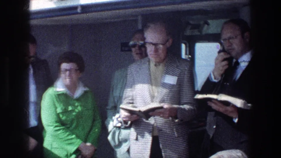 On a 1976 Sea of Galilee boat, a man in a tan jacket reads from a black book beside other adult travelers.