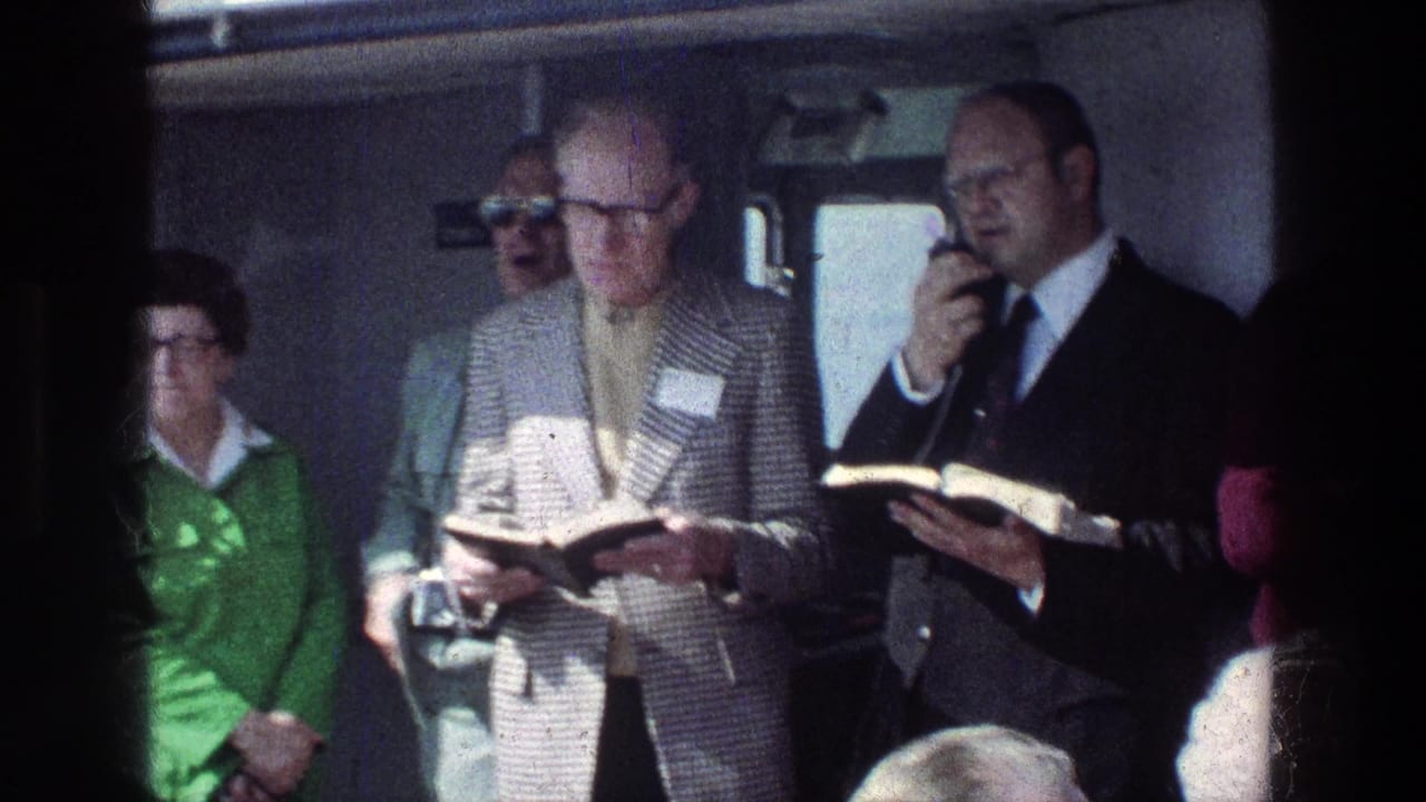 Tour passengers stand inside a vehicle while two men hold open books near a microphone.