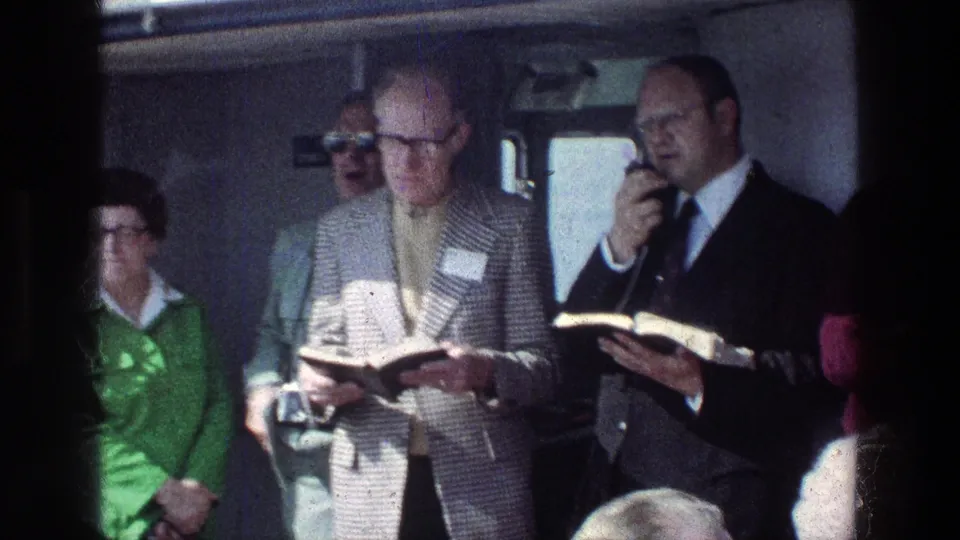 1976 Sea of Galilee tour scene with passengers inside a vehicle as a suited man speaks into a microphone beside another man holding a book.