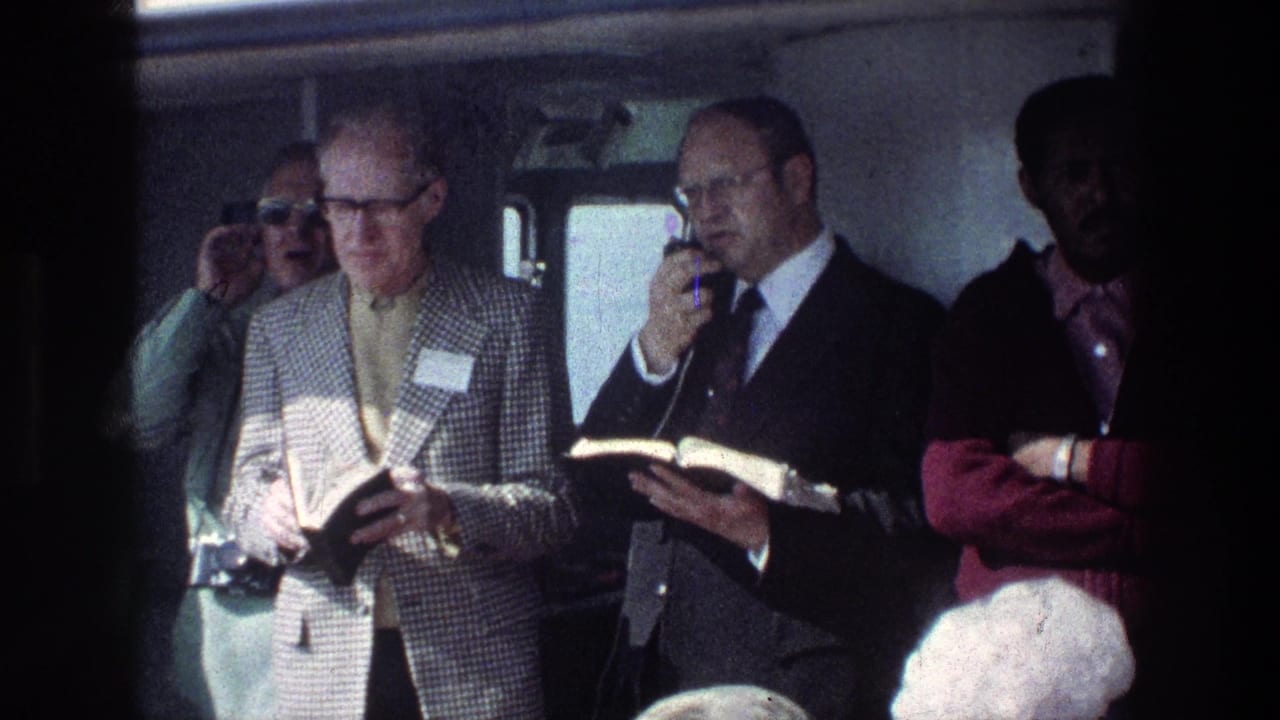 A suited man speaks into a microphone while passengers stand around him with books.