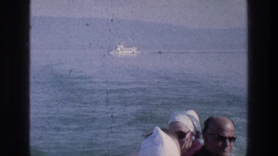 Sea of Galilee view from the tour setting, with a small distant boat on calm water and passengers partly visible in the foreground.
