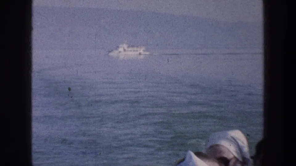 1976 Sea of Galilee home-movie frame with a white passenger boat crossing blue water beyond dark window framing.