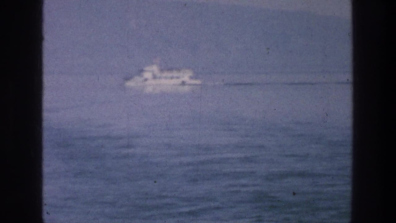 A white passenger boat passes across the Sea of Galilee while partial observers appear at the bottom of the 8mm frame.