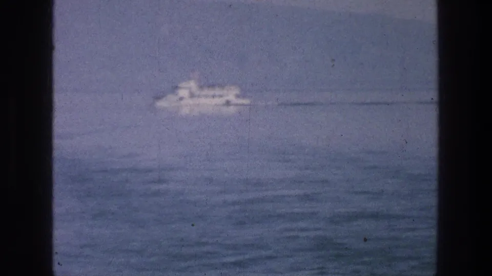 A white passenger boat passes across the Sea of Galilee while partial observers appear at the bottom of the 8mm frame.