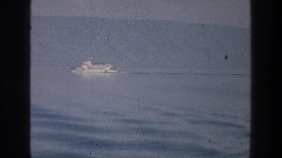 1976 Sea of Galilee home-movie still with a white passenger boat crossing calm water below hazy hills.