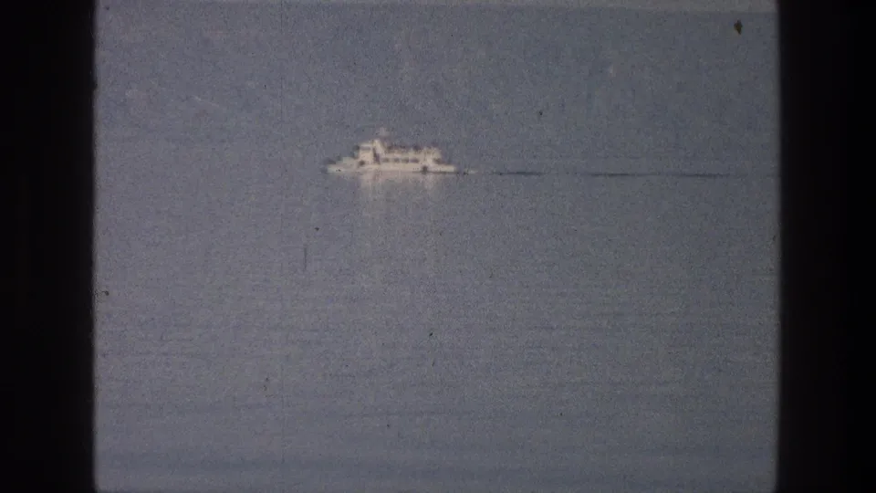 Small white boat on the Sea of Galilee in 1976-era 8mm footage, seen at a distance across muted blue-gray water.