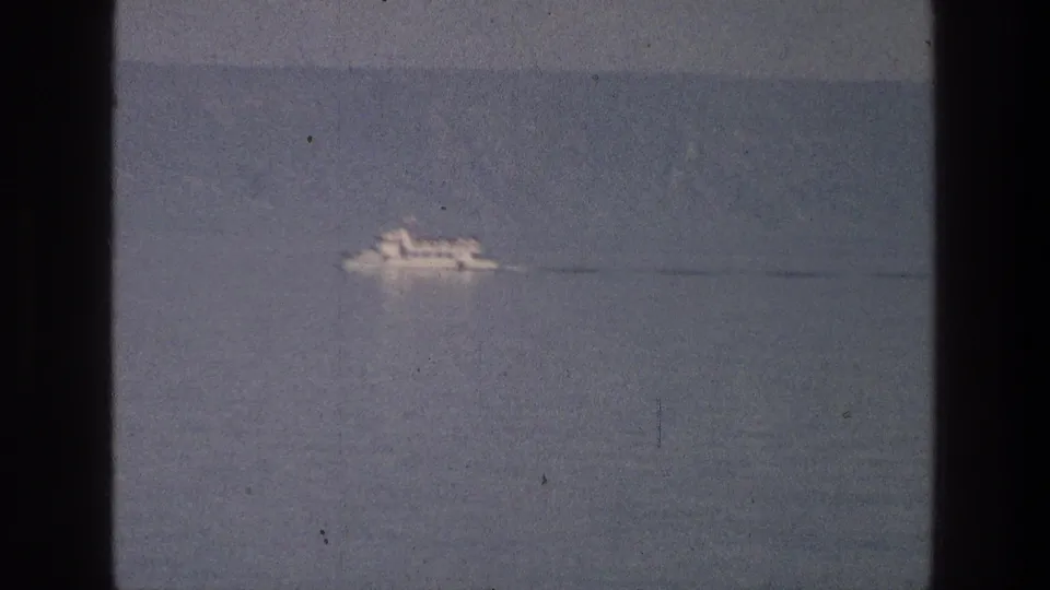 The white boat remains distant on the Sea of Galilee, moving across the muted lake under a hazy overcast sky.