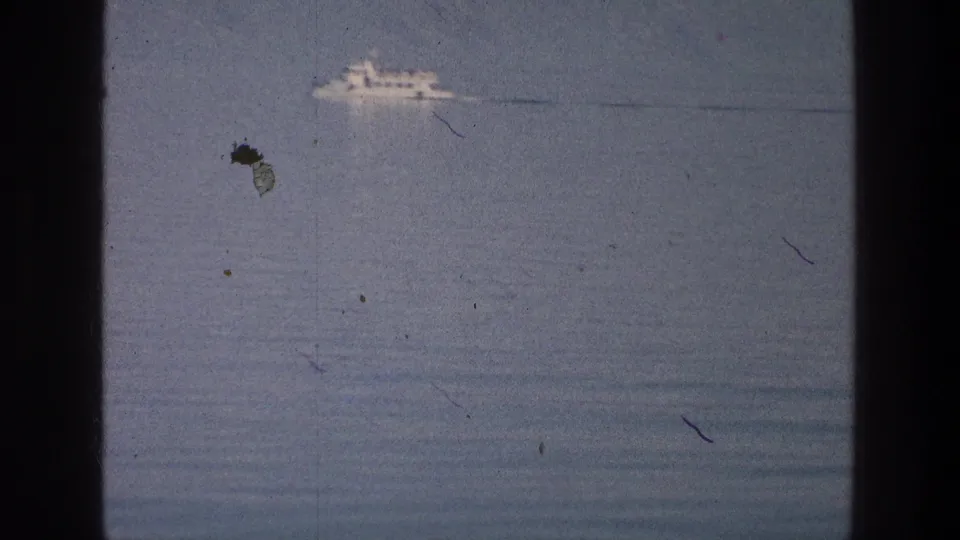 1976 Sea of Galilee home-movie frame showing a distant white tour boat and wake, with visible 8mm dust and scratches.