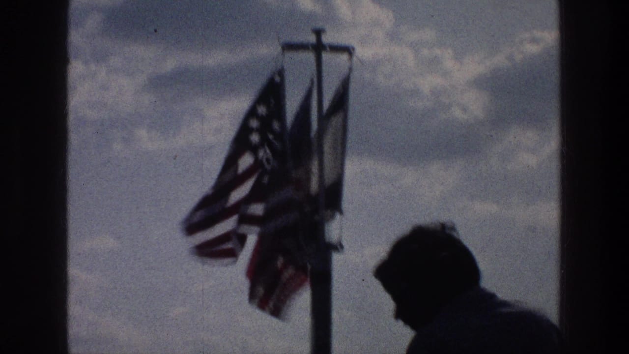Silhouetted young man near a flagpole with flags against cloudy sky and distant water.