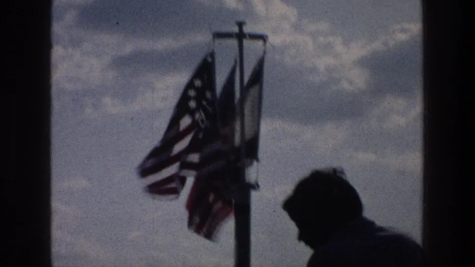 1976 Sea of Galilee home-movie frame showing a silhouetted young man beneath flags on a pole, with cloudy sky and distant water behind.