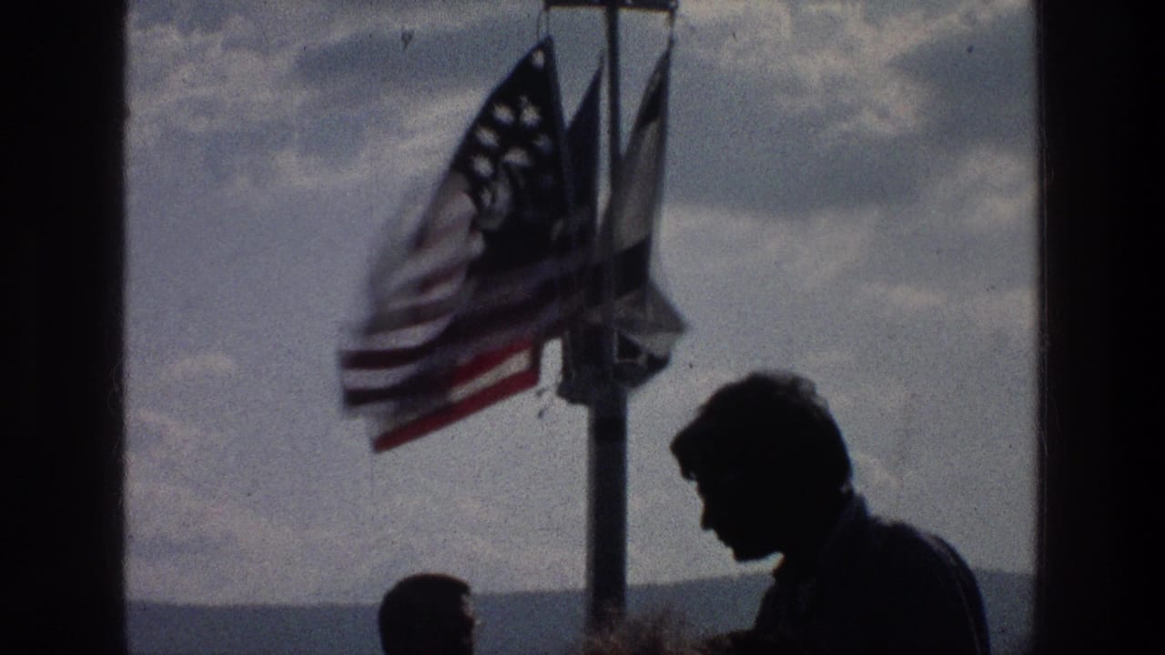 1976 Sea of Galilee frame with two silhouetted figures near a pole flying an American flag and two other banners under a cloudy sky.