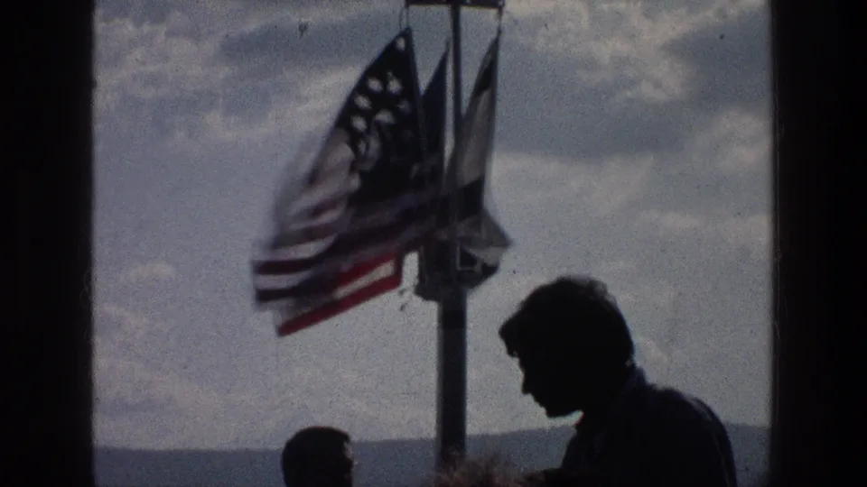 1976 Sea of Galilee frame with two silhouetted figures near a pole flying an American flag and two other banners under a cloudy sky.