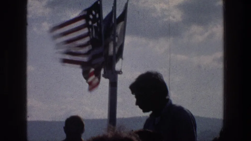 1976 Sea of Galilee frame showing a young man in silhouette, another figure lower in frame, and flags blowing on a pole.