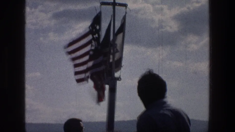 1976 Sea of Galilee home-movie frame with a silhouetted man facing flags, including an American flag, above distant hills and water.