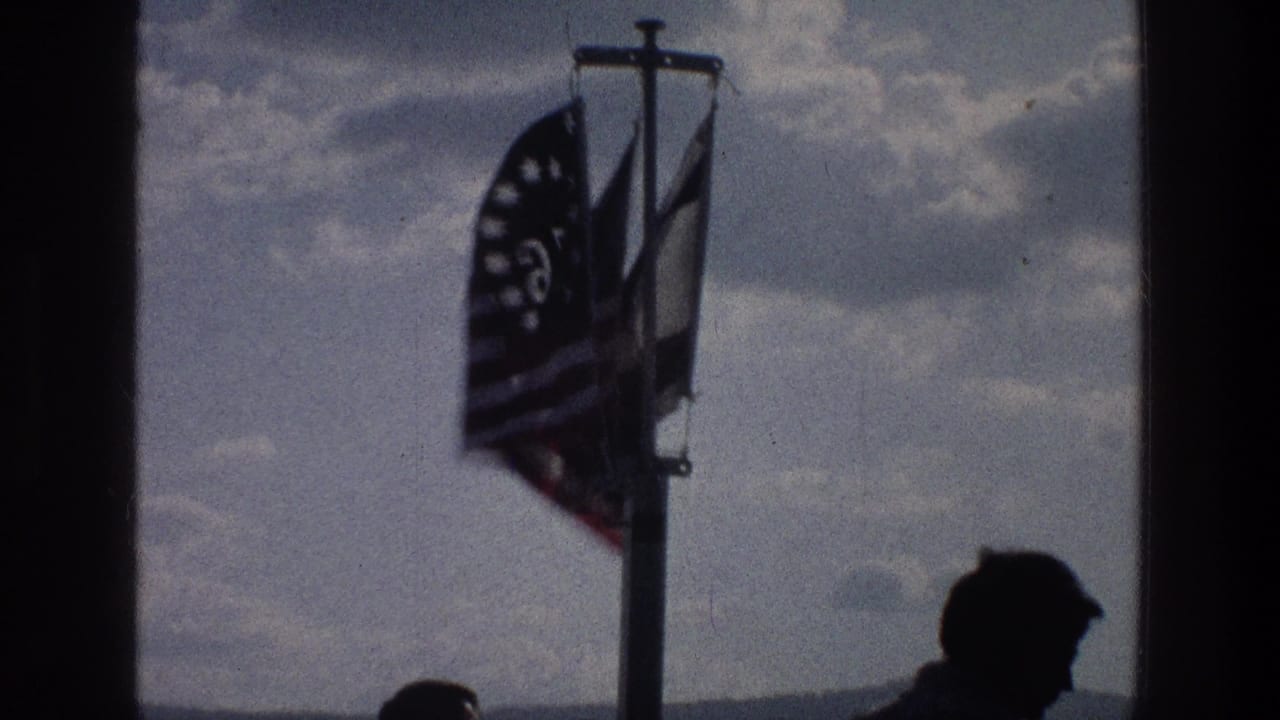 A 1976 Sea of Galilee home-movie frame shows flags in wind beside a dark vertical divider and a silhouetted figure below.