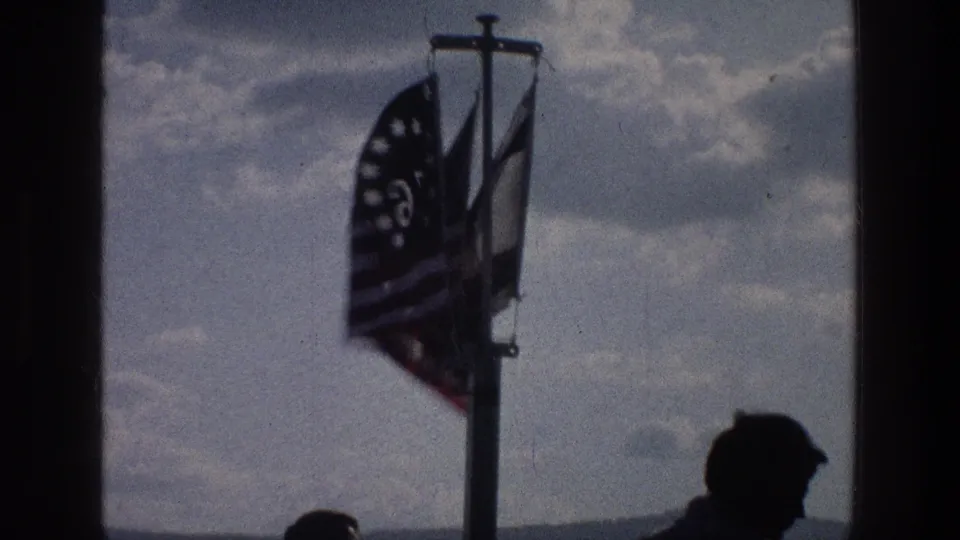 A 1976 Sea of Galilee home-movie frame shows flags in wind beside a dark vertical divider and a silhouetted figure below.