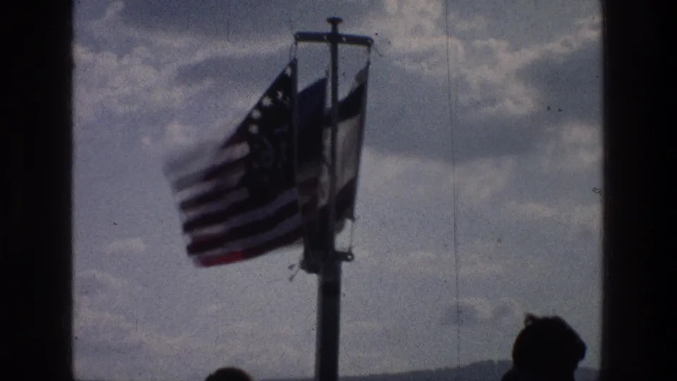 A 1976 Sea of Galilee frame shows three flags on one pole under a cloudy sky, framed by dark edges and a silhouetted viewer.