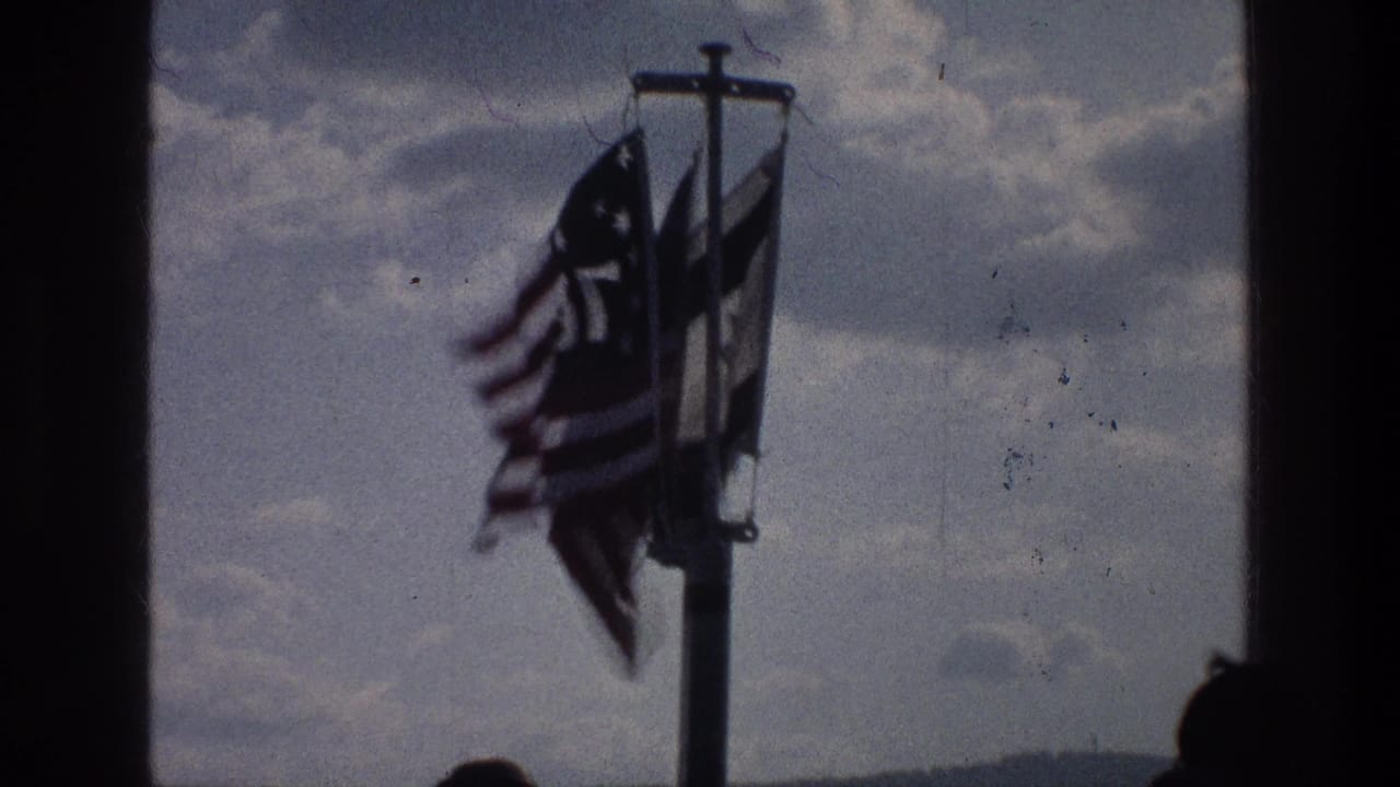 Flags on a boat pole seen through a dark window frame, with sky and partial passenger heads below.