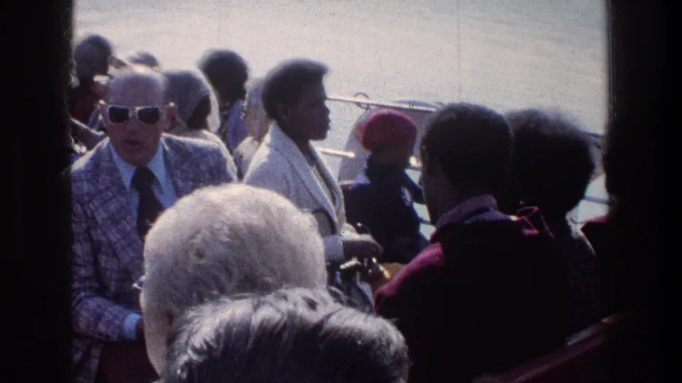 A handheld 1976 boat view shows passengers seated close together, with an older man in sunglasses and figures turned toward the water.
