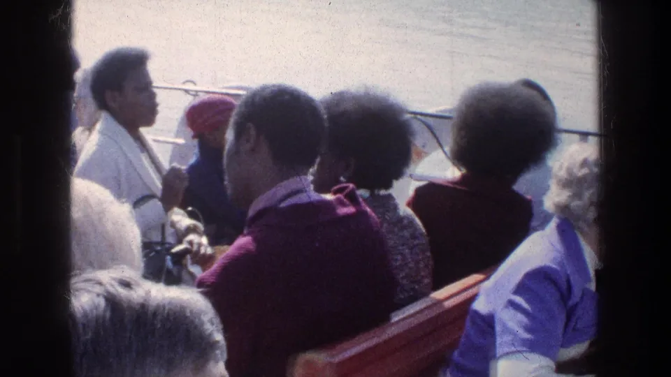 Passengers aboard a 1976 Sea of Galilee boat face the rail and one another, with red seats and bright water in the background.