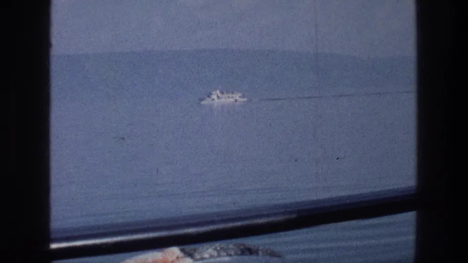 Distant white vessel on the Sea of Galilee in 1976, seen through a window with soft 8mm color and a faint wake trailing behind it.