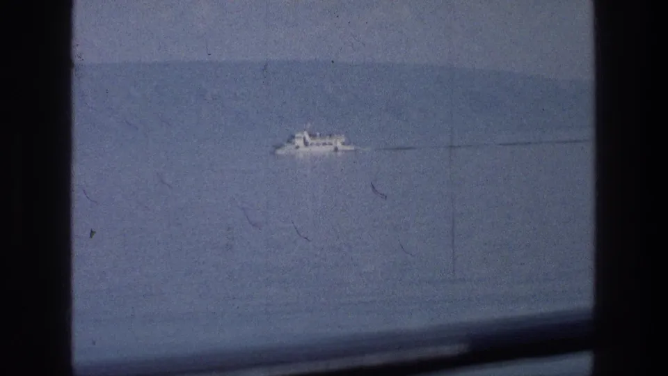 1976 home-movie frame of a white boat moving across the Sea of Galilee, partially framed by dark window bars and faded blue film tones.
