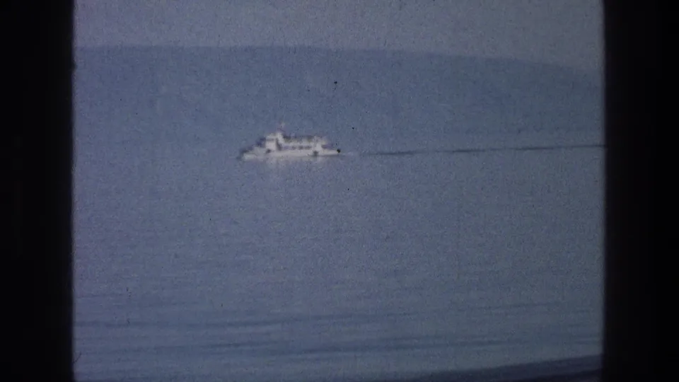 A distant white boat leaves a faint wake on the Sea of Galilee in this 1976 8mm travel view through a dark window frame.