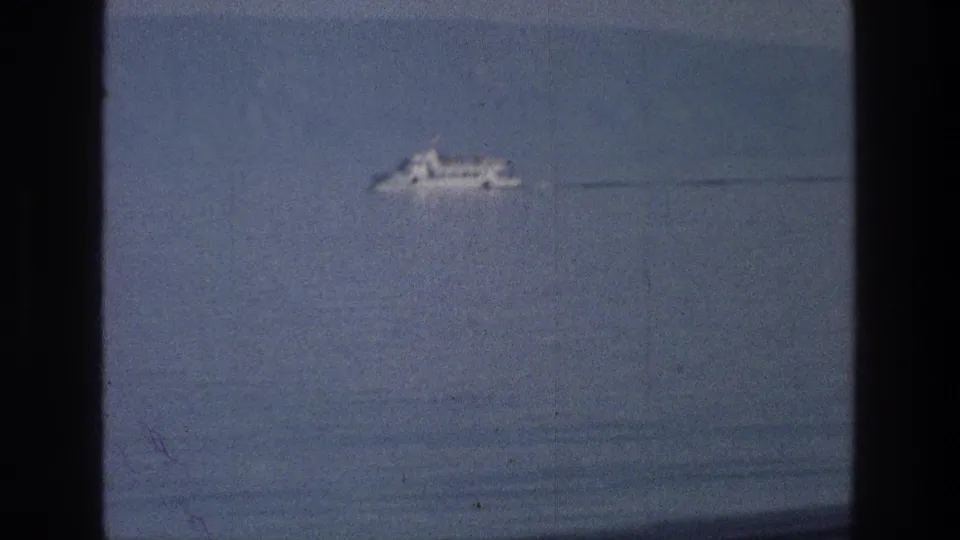1976 8mm Sea of Galilee frame showing a distant white boat on calm blue water with soft focus and dark gate-edge borders.