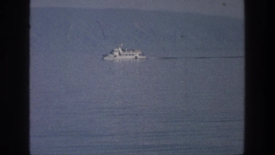 The white vessel appears farther across the Sea of Galilee, leaving a faint wake within a grainy 1970s 8mm frame.