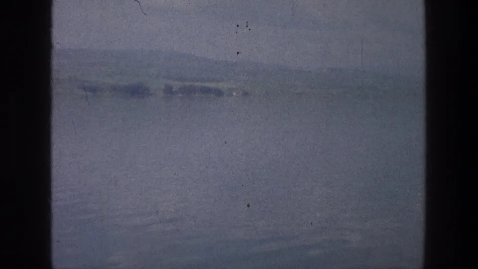 A soft 8mm view of the Sea of Galilee shows calm water and a hazy low shoreline after the camera pans from the boat.